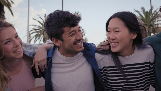Close up multiracial group young friends standing hugging urban street palm trees. Millennial people laughing having fun outdoors on university vacation. Concept cheerful students embracing together. 
