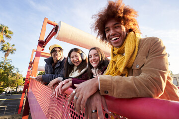 Multi-ethnic group smiling friends gathered on street leaning on railing. Happy young people enjoying together outdoors pose for portrait. Social relationships in college students of Generation z. - obrazy, fototapety, plakaty