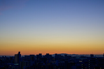 都市の夜明け。日の出とともに空と雲がオレンジ色に染まり、ビル群はシルエットとして写す。神戸市東灘区から大阪方面をのぞむ