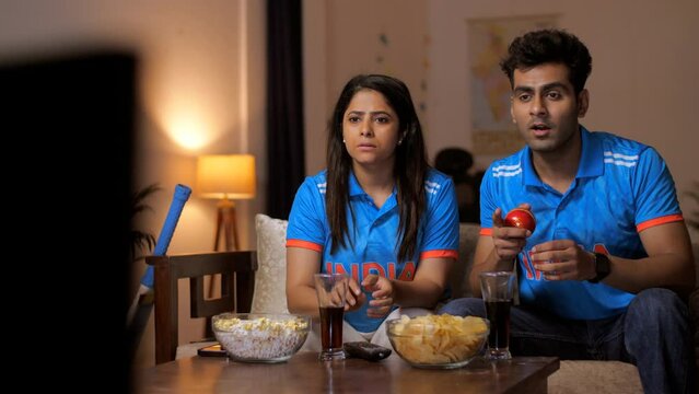 Friends in Indian jerseys sitting on the couch and watching a live cricket match on TV/ television - cricket fever, World Cup 2023. Indian cricket fan wearing an Indian jersey - excited  cheering 