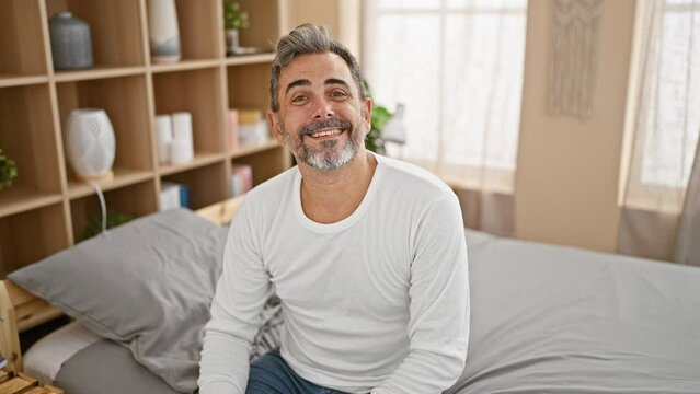 Cheerful, confident young hispanic grey-haired man enjoying a relaxed morning in bed, happily taking off his glasses while sitting comfortably in his cozy bedroom