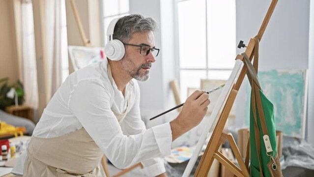 At the studio, a relaxed, young hispanic man with grey-haired beard, artist and student, drawn to music, drawing away on canvas, headphones on, bringing art to life indoors