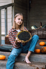 Girl is sitting on steps of old village house holding large sunflower in hands. Ripe pumpkins and baskets filled with harvested crops: apples and zucchini lie nearby. Harvest. Autumn. Thanksgiving day