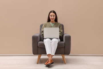 Fototapeta na wymiar Beautiful woman with laptop sitting in armchair near beige wall indoors