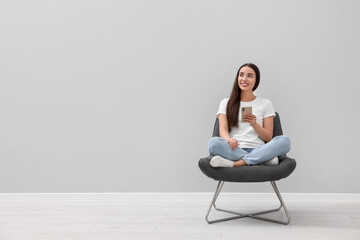 Beautiful woman with smartphone sitting in armchair near light grey wall indoors, space for text