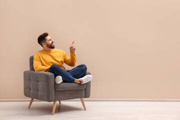 Handsome man pointing at something while sitting in armchair near beige wall indoors, space for text