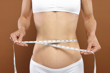 Slim woman measuring waist with tape on brown background, closeup. Weight loss