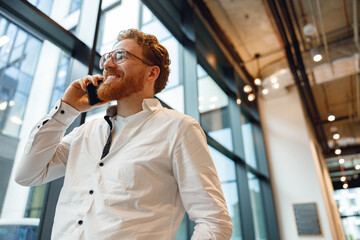 Smiling male manager in eyeglasses is talking phone standing in modern coworking space