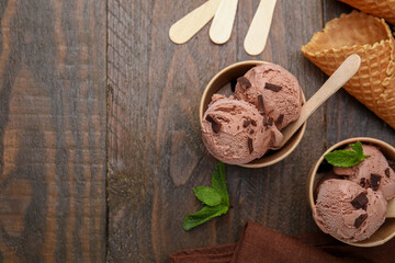 Paper cups with tasty chocolate ice cream, sticks, mint leaves and cones on wooden table, flat lay....