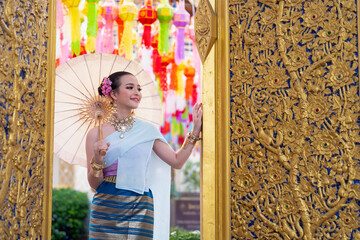 Asian woman wearing a traditional Lanna dress smiled happy and holding paper Thai umbrella in front of a golden carved wooden door during Yi Peng lantern festival. Lamphun Province, Thailand.