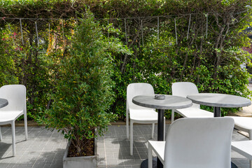 Empty tables with chairs against a background of green vegetation in a street cafe. There is an empty metal ashtray on the table.