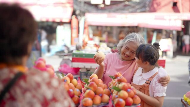 Happy Asian family grandmother and grandchild girl choosing and buying fresh fruit together at street market. Senior woman and little girl enjoy outdoor lifestyle travel in the city on summer vacation