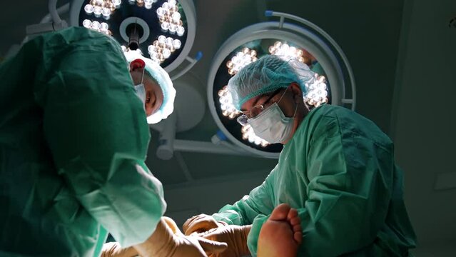 Younger surgeon holds patient's foot and older surgeon applies tool. Low angle view on the doctors performing surgery.