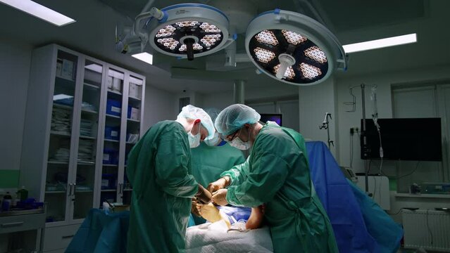 Careful surgeons operate patient's foot. Doctors apply tools with precaution. Two bright round lamps light the surgery room.