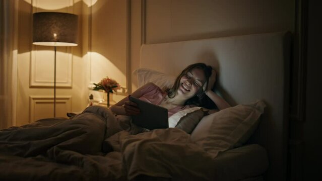Girl watching funny tablet video movie at night. Happy woman resting home bed