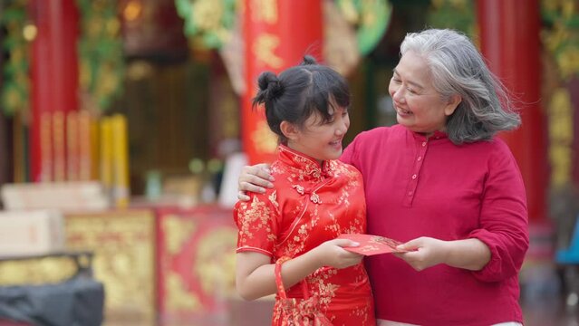 4K Happy Asian family grandmother giving red envelope contained money gift with blessing to little grandchild girl in Chinese red dress. Chinese lunar new year festival and holiday celebration concept