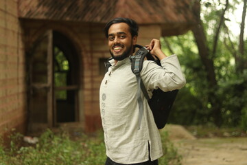 Fototapeta na wymiar An Indian boy with black beard wearing khadi kurta and having a backpack with green vegetation in background and lifting the bag with a smile