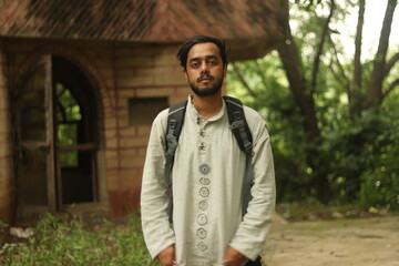 An Indian boy with black beard wearing khadi kurta and having a backpack with green vegetation in background and standing straight