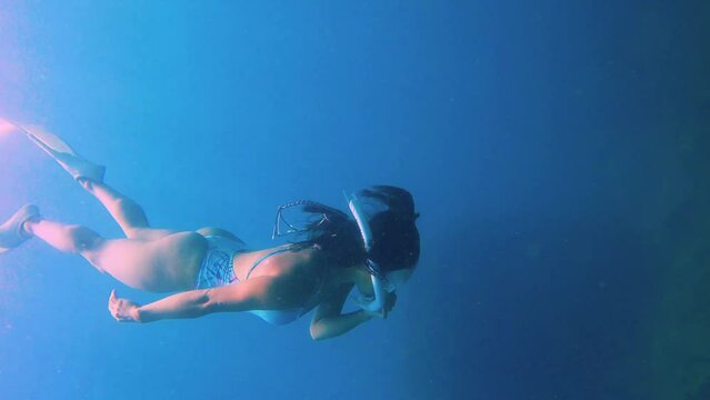side view to back close up to a young asian female free diver wearing long fins white swim suit and snorkel mask duck diving down at bottom sea floor seabed with coral rocks in low visibility ocean