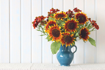decorative sunflowers and helium in a blue jug on a table against a white board wall. Garden summer flowers.
