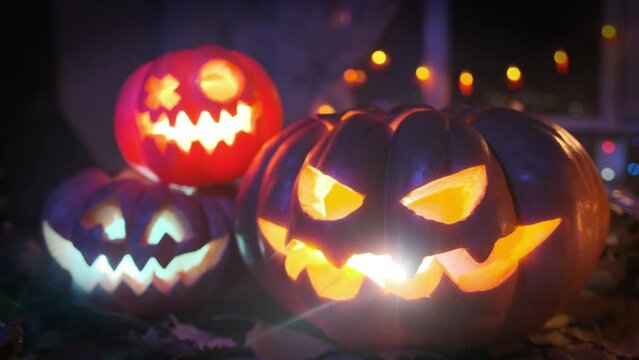 Three holiday lanterns made of carved pumpkins, glow with multi-colored lights in the smoke in the dark near house among cones dry leaves and colorful garlands. Halloween background