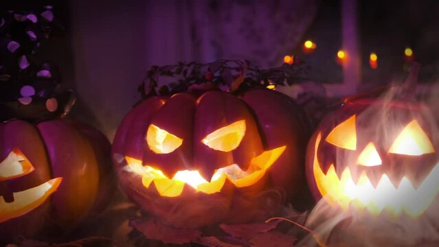Many carved pumpkin jack o lanterns stand on table and glow with colorful lights in smoke against the backdrop of garlands. Halloween background tracking shot.