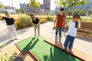 Cute school girl playing mini golf with family. Happy toddler child having fun with outdoor activity. Summer sport for children and adults, outdoors. Family vacations or resort.