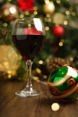 Glass of red wine on wooden table with Christmas decorations. Christmas tree and lights in the background.
