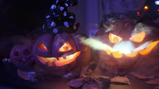 Tracking shot close up burning pumpkins stand in row among dry leaves, flickering candles, garlands, jack-o'-lantern in with hat lying on threshold in fog celebrating Halloween background.