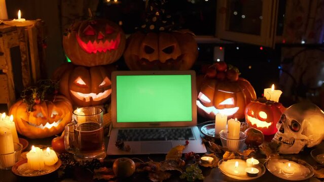 Flashing Carved pumpkins with open laptop with green screen next to beer mug holiday background cozy workplace with halloween decorations, candles and garlands.
