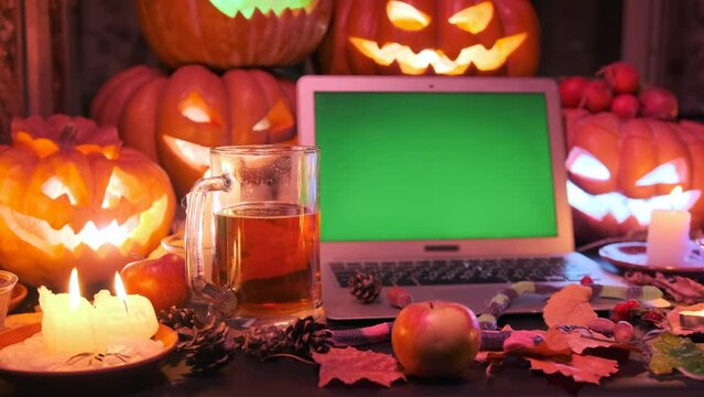 Halloween time background, laptop with chromakey green screen, glowing carved pumpkins and glass of beer,  candles, dry leaves, in dark.
