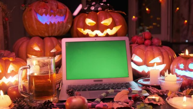 Laptop with chromakey screen, pile pumpkins lanterns, candles and mug beer, slowly shot. Creepy Happy Halloween party celebration mystical background. Frightening invitation. Close up.