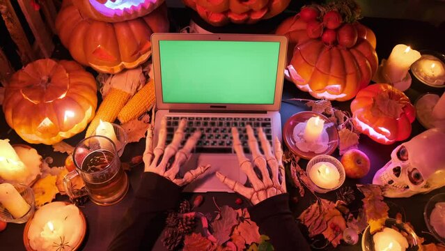 Top view of bony monster hands typing at laptop keyboard with green screen with many cut out orange traditional pumpkins, candles, skull and mug beer. Festive Halloween eve background.
