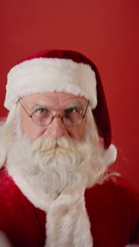 Close-up vertical shot of funky Santa Claus in red coat, hat and glasses, with white beard and moustache dancing energetically with batman moves, on red background. Template, copy space