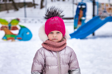 Sad little child girl in red hat posing on winter snowy playground, bored looking at camera. Portrait of adorable kid walk of winter holiday. Concept of wintertime and childhood. Copy ad text space