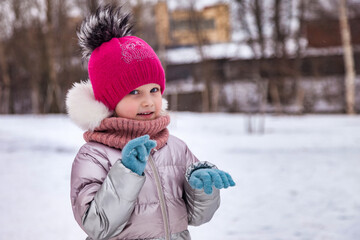 Portrait of funny child girl posing in winter urban public park in warm clothing, smiling looking at camera. Little kid walking winter holiday. Concept of wintertime and childhood. Copy ad text space