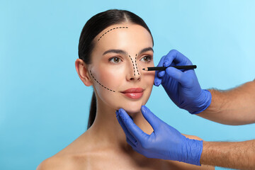 Woman preparing for cosmetic surgery, light blue background. Doctor drawing markings on her face,...