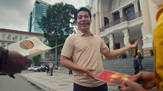 Medium shot of smiling Asian male travel guide standing in front of Opera House in Ho Chi Minh city and conducting sightseeing tour for unrecognizable diverse couple holding tourist brochures