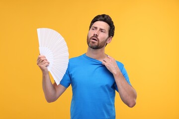 Unhappy man with hand fan suffering from heat on orange background