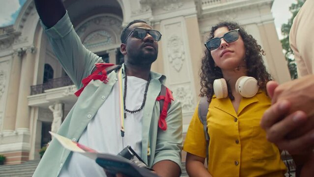 Medium shot of African American male tourist and Caucaisan wife standing in square in Ho Chi Minh city next to Opera House, asking guide question, pointing to landmark in tour booklet, then away