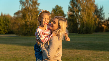 Happy mother holds laughing little daughter in autumn park at sunset. Child with mother outdoors. Weekend, leisure, active games