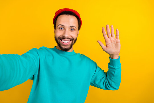 Portrait of optimistic nice man with beard dressed turquoise pullover make selfie waving palm say hi isolated on yellow color background