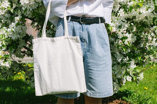 Woman hand holding eco bag. Female hold white canvas textille mesh zero waste shopper on city street, green apple tree flowers background. Blank mock up tote bag, place for text design logo. 