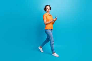 Fototapeta na wymiar Full size photo of smart girl dressed stylish polo denim pants walk in empty space with smartphone isolated on blue color background