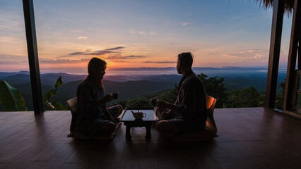 a couple of men and women on vacation in Northern Thailand , staying at a homestay cabin hut in the mountains of Chiang Rai Doi Chang drinking tea on the wooden balcony at sunrise - obrazy, fototapety, plakaty