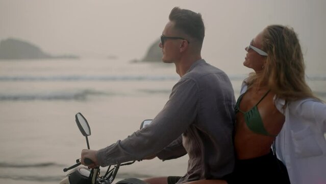 Young couple riding motorcycle on ocean beach at sunrise. Man, woman travelling by motor bike on coastline along sea. Lady looks back to camera, laughs, spreads arms, touches hair blowing in the wind.
