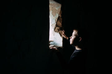 A little boy in a black T-shirt is standing in a house near a window. The child in the room is looking at the old window from which the light is coming. Consequences of war