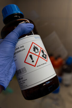 Hand with a blue gloves holding glass bottle with an organic solvent. Glassware with hazardous symbol in the chemical lab.