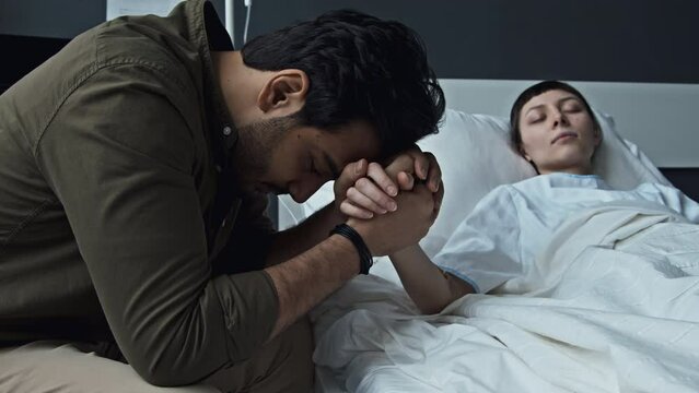 Selective focus medium close-up shot of worried Middle Eastern man holding hand of his beloved lying in hospital bed in coma