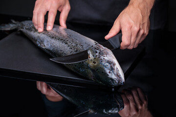 A cook on a black background cuts a whole salmon carcass.